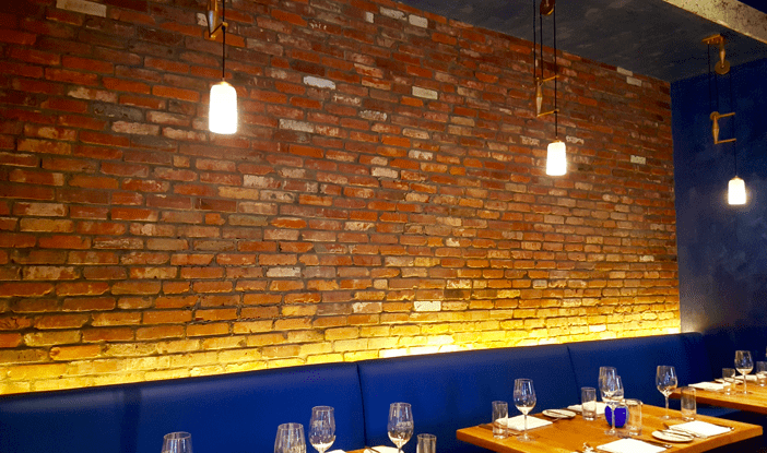 photo of a thin brick wall above a long blue banquette, with empty set tables.