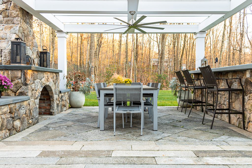 photo of light gray  and tan paving planks in a beautiful outdoor bar area.  A fireplace is on one side, and a stone bar and high top chairs is on the other side, with a gray table in between.