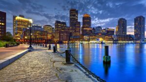 A cobblestone path bordering a bay, with a city in the background, at sunset.