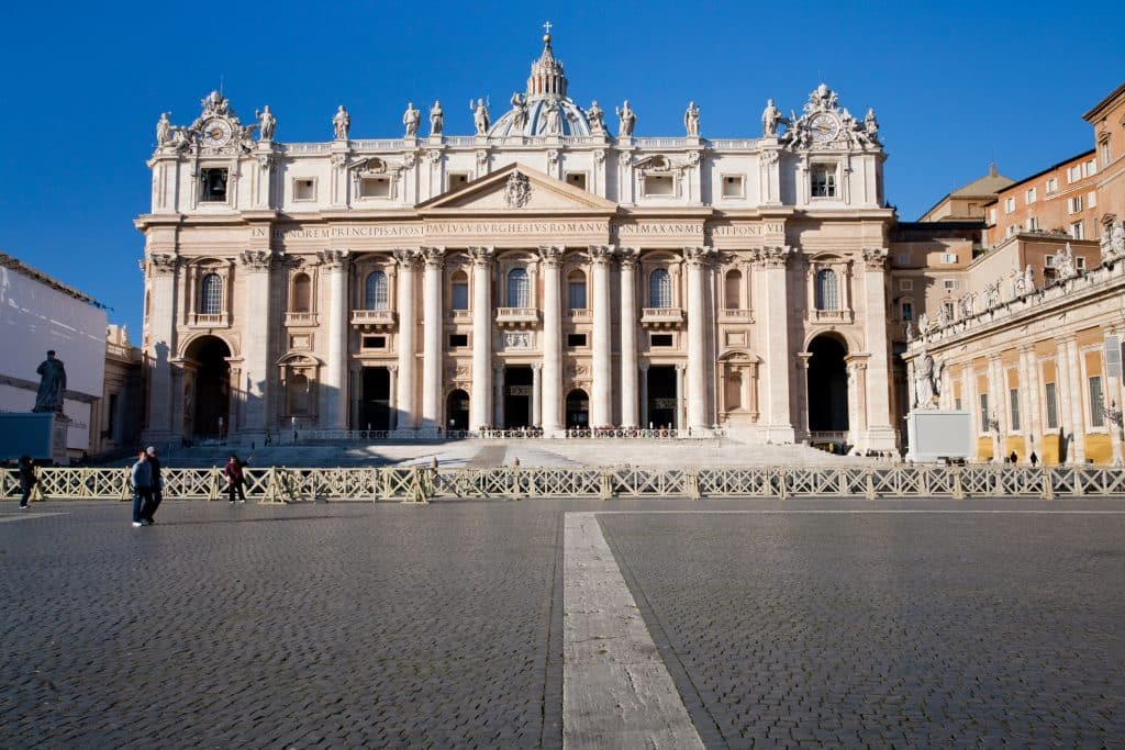 A photograph of St. Peter's Square, in Vatican City.  The Basilica is in the background. Sampietrini, a small, dark European cobblestone, cover the grounds of the Square in the foreground.