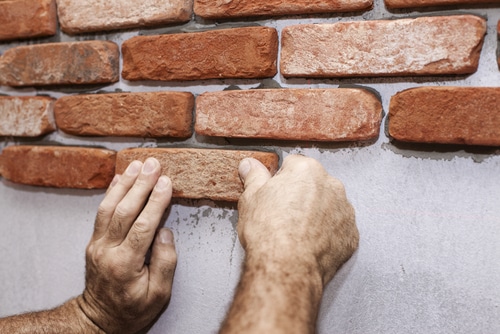 close-up photo of a man's hands as he's setting thin brick veneer into mortar on a wall.