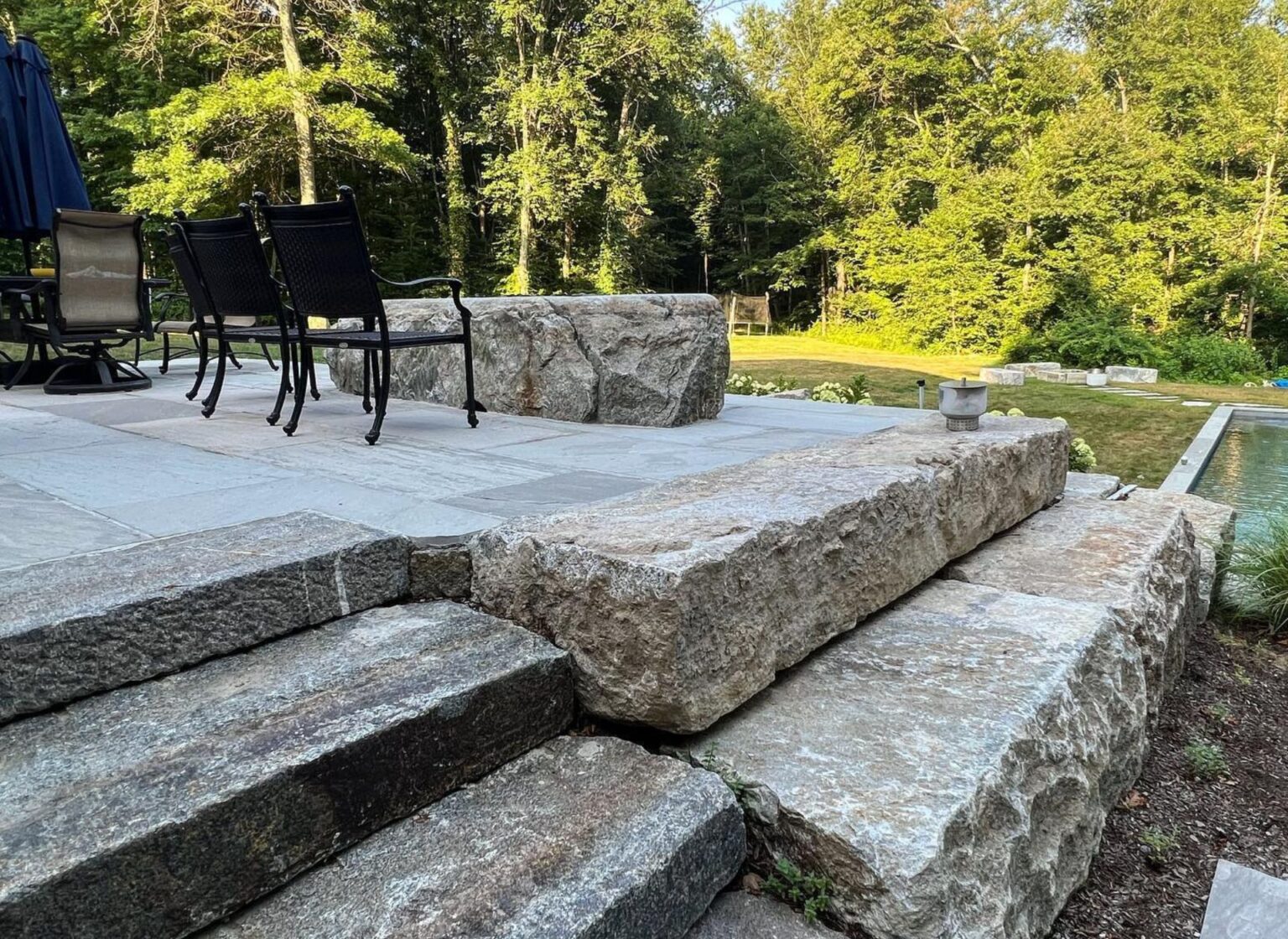 A set of large reclaimed granite curbing blocks forming broad steps that lead from a lower patio up to a raised stone terrace with outdoor seating, set against a backdrop of dense green woods.
