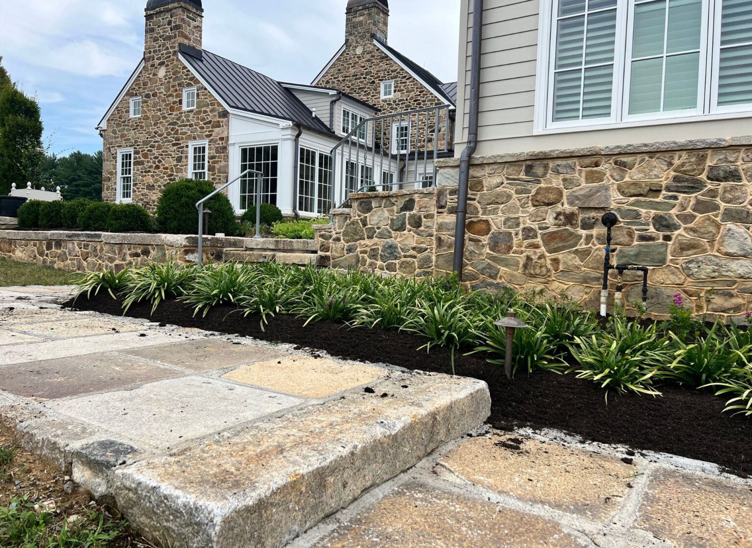 A reclaimed granite step walkway made from large weathered stone slabs running beside a stone‑faced house, bordered by fresh mulch and low green plantings.