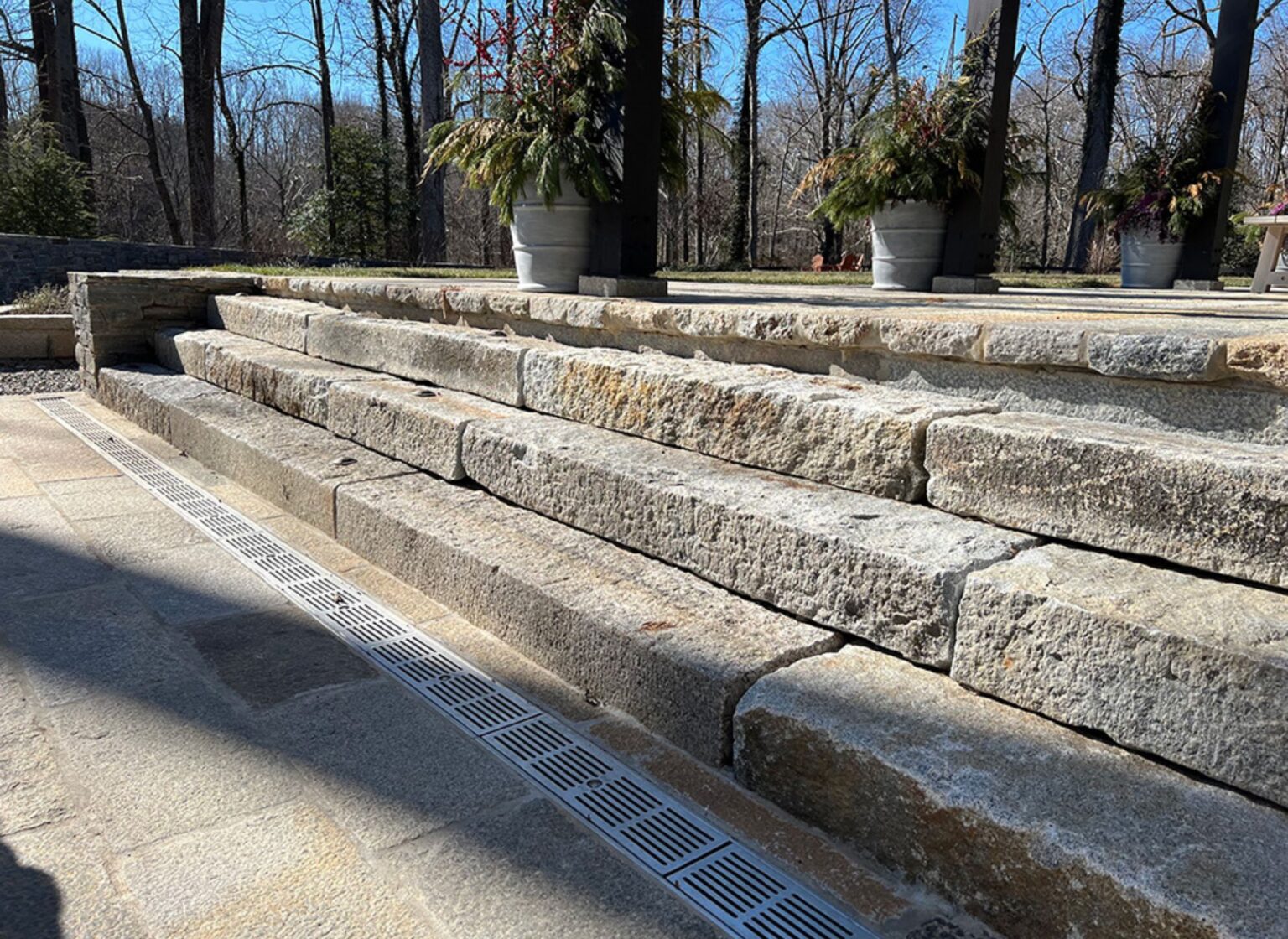 A series of wide antique stone patio steps built from thick, weathered stone blocks, leading up to a raised terrace bordered by planters and surrounded by tall trees.