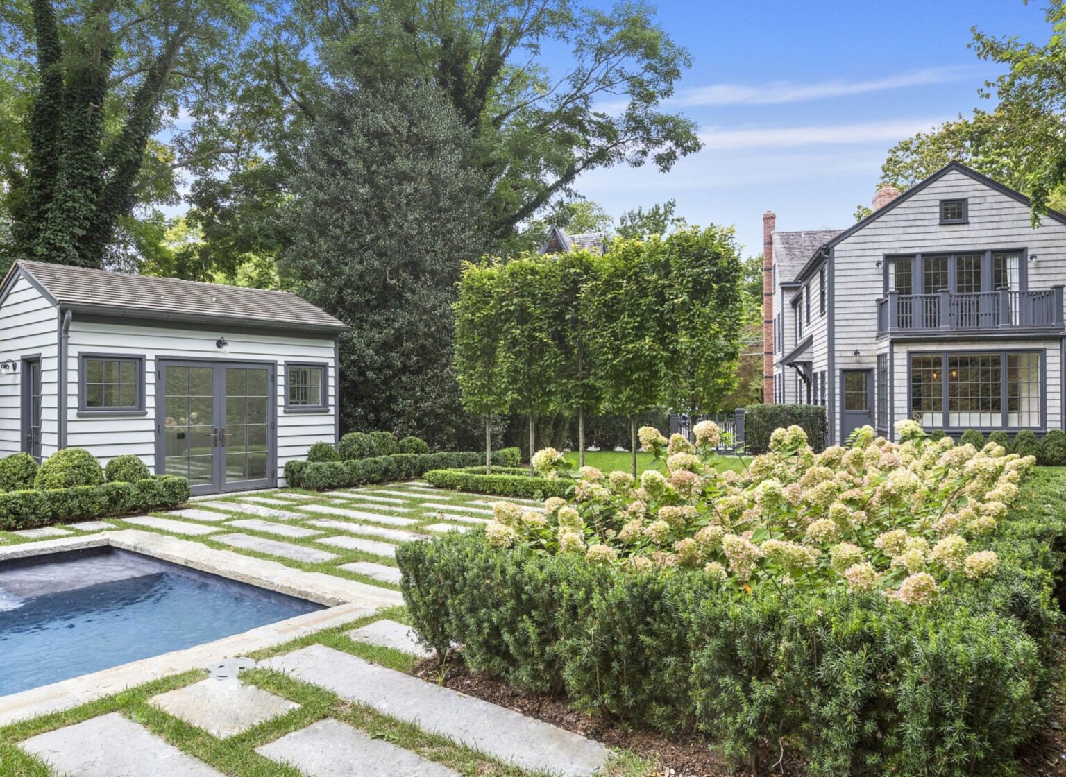 A rectangular swimming pool finished with antique granite curbing pool coping, bordered by a manicured garden with stone walkways and set beside a traditional clapboard home.