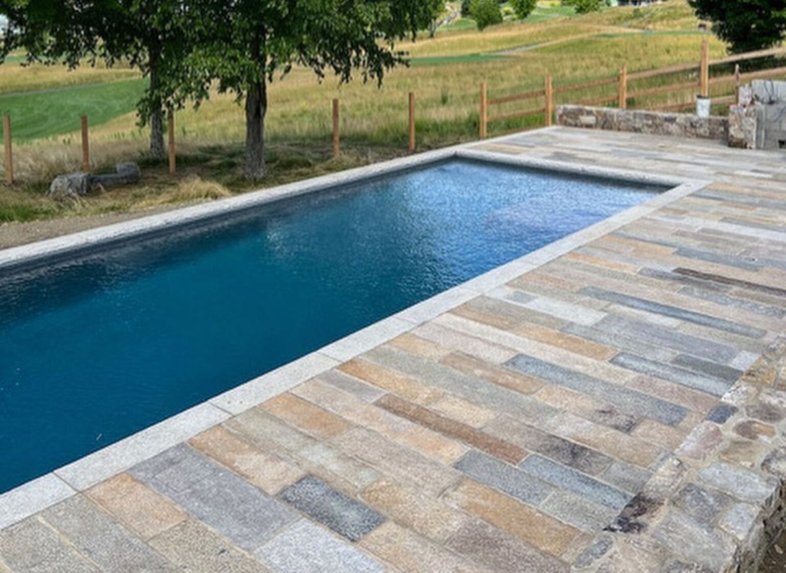 A rectangular swimming pool finished with antique granite plank pool coping, featuring long weathered stone planks surrounding the water and set within a rural landscape with fencing, trees, and open fields.