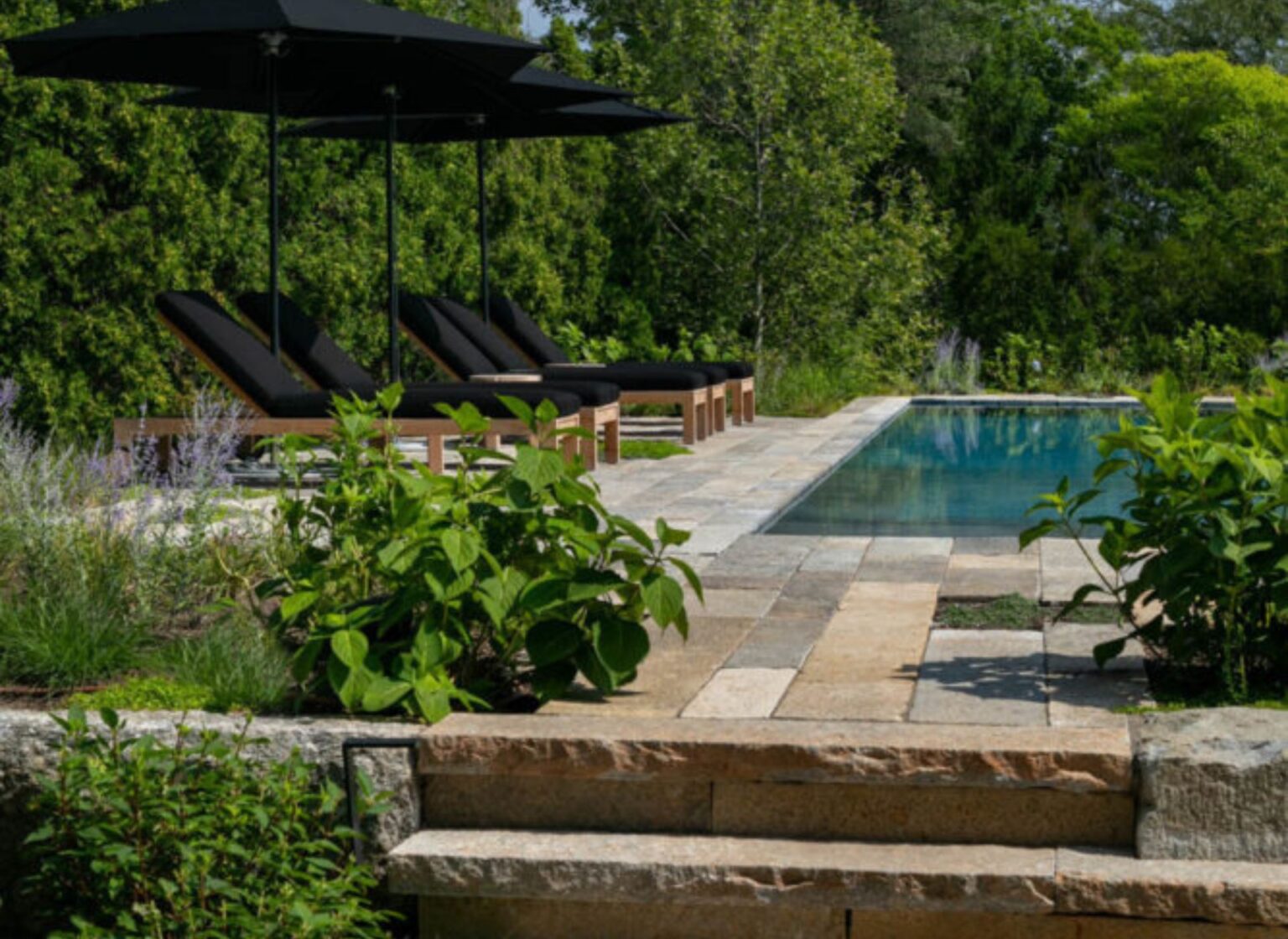 A rectangular swimming pool surrounded by an antique granite plank pool deck, with long weathered stone planks forming a patio area, furnished with lounge seating and umbrellas, set within dense greenery.