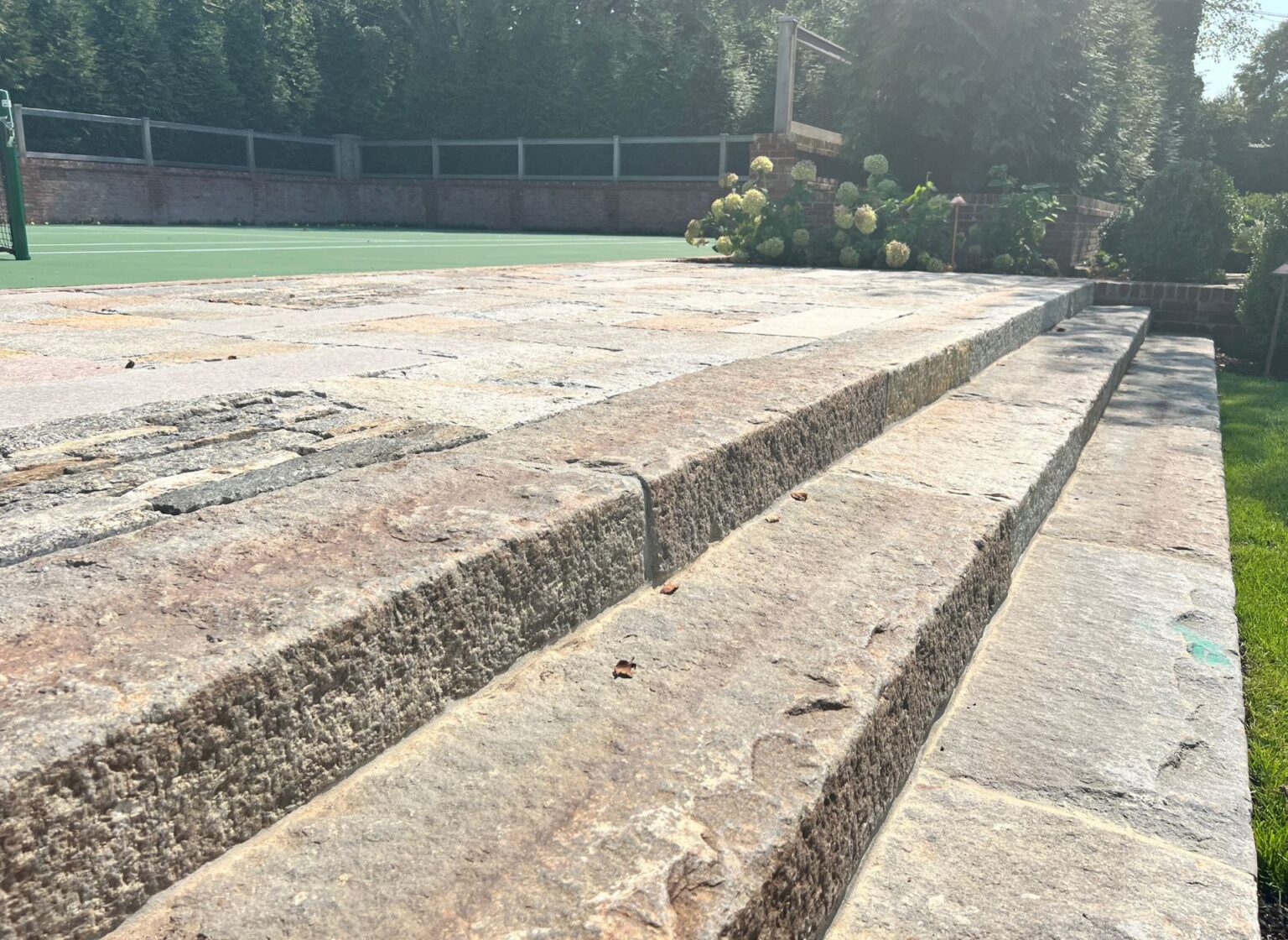 A set of antique granite curb steps with rough textured faces leading up to a sports court, bordered by a lawn and surrounded by mature landscaping.