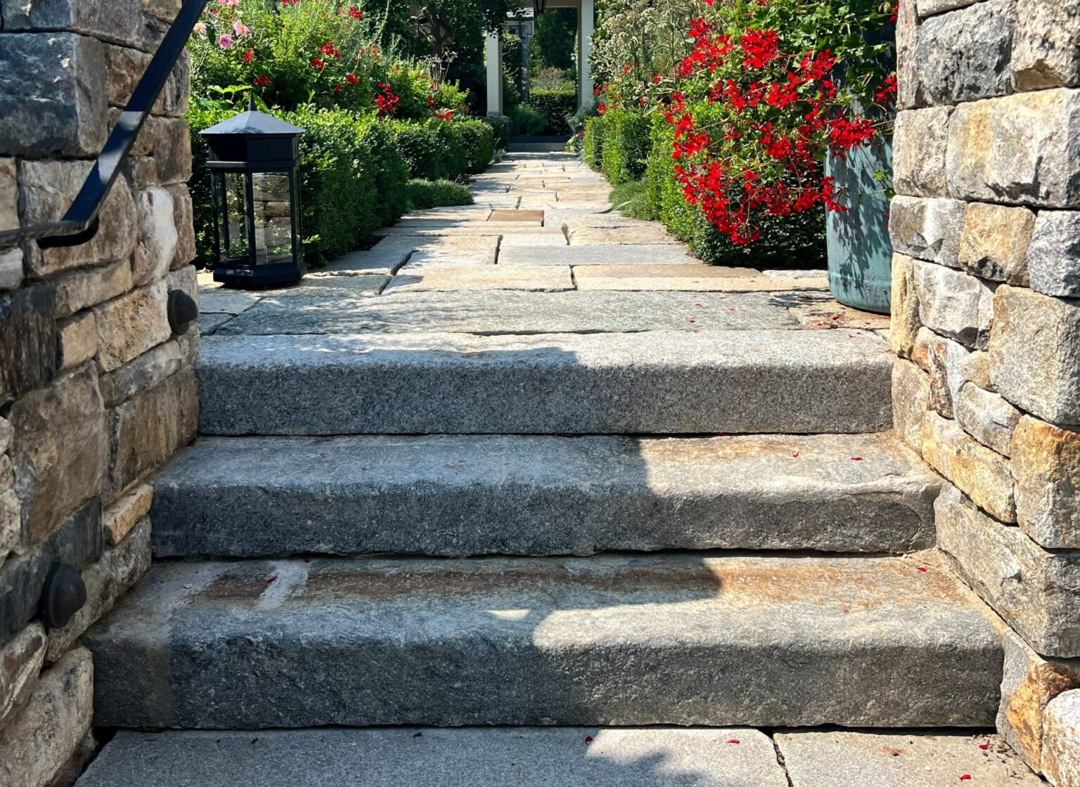 A series of reclaimed granite curbing steps leading from a lower stone patio up toward a garden walkway, framed by stone walls and vibrant red flowering plants.