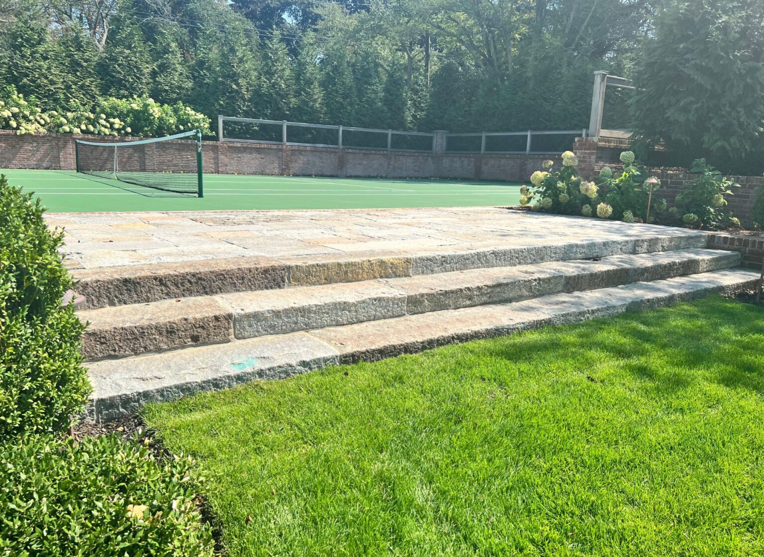 A wide set of reclaimed granite steps leading from a lawn up to a tennis court, with neatly trimmed shrubs and trees bordering the area.