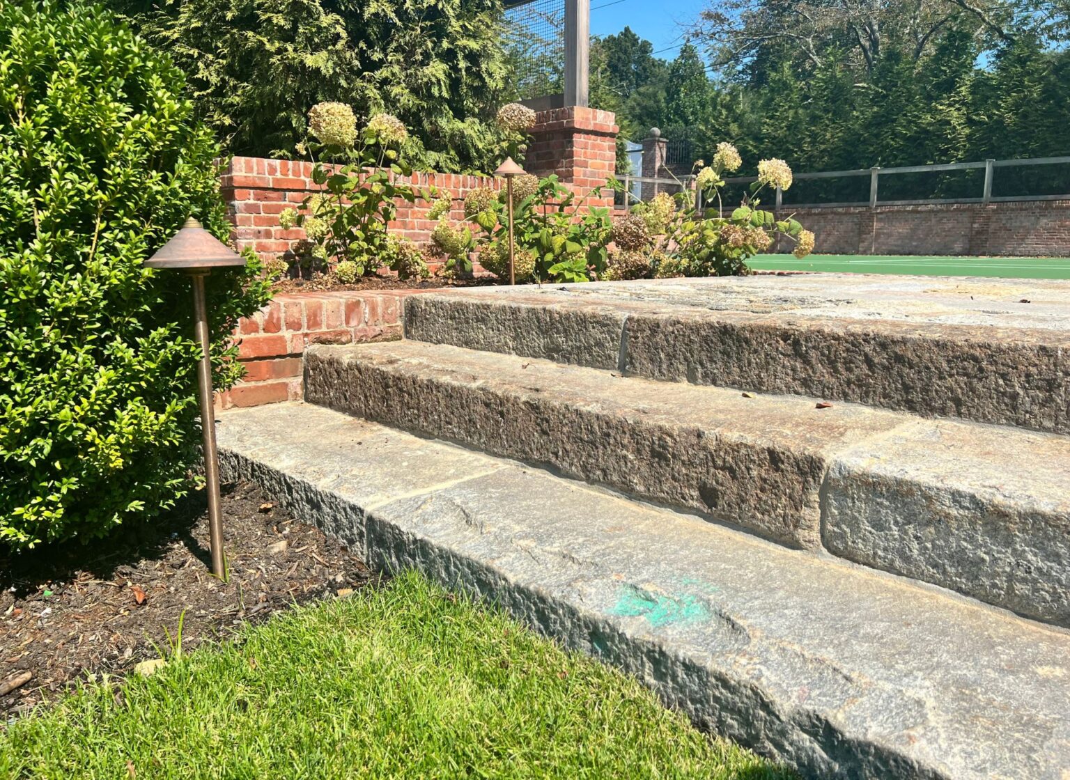 Wide outdoor steps built from thick reclaimed granite treads, rising alongside a reclaimed brick landscape wall at the edge of a tennis court, bordered by lawn and garden plantings.