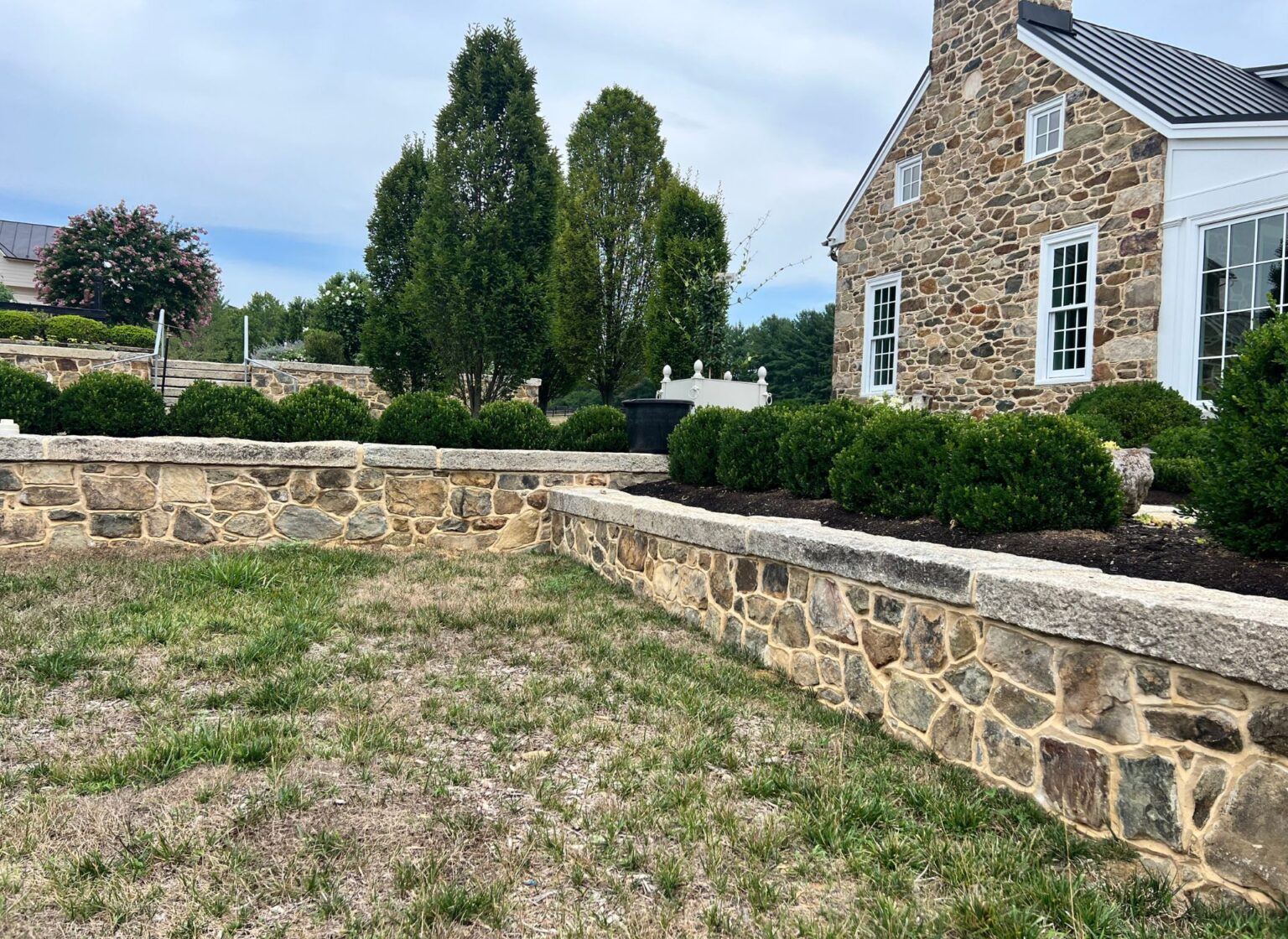 A low landscape retaining wall built from reclaimed fieldstone, topped with thick stone wall caps and running across a sloped lawn in front of a stone‑faced home with manicured shrubs.