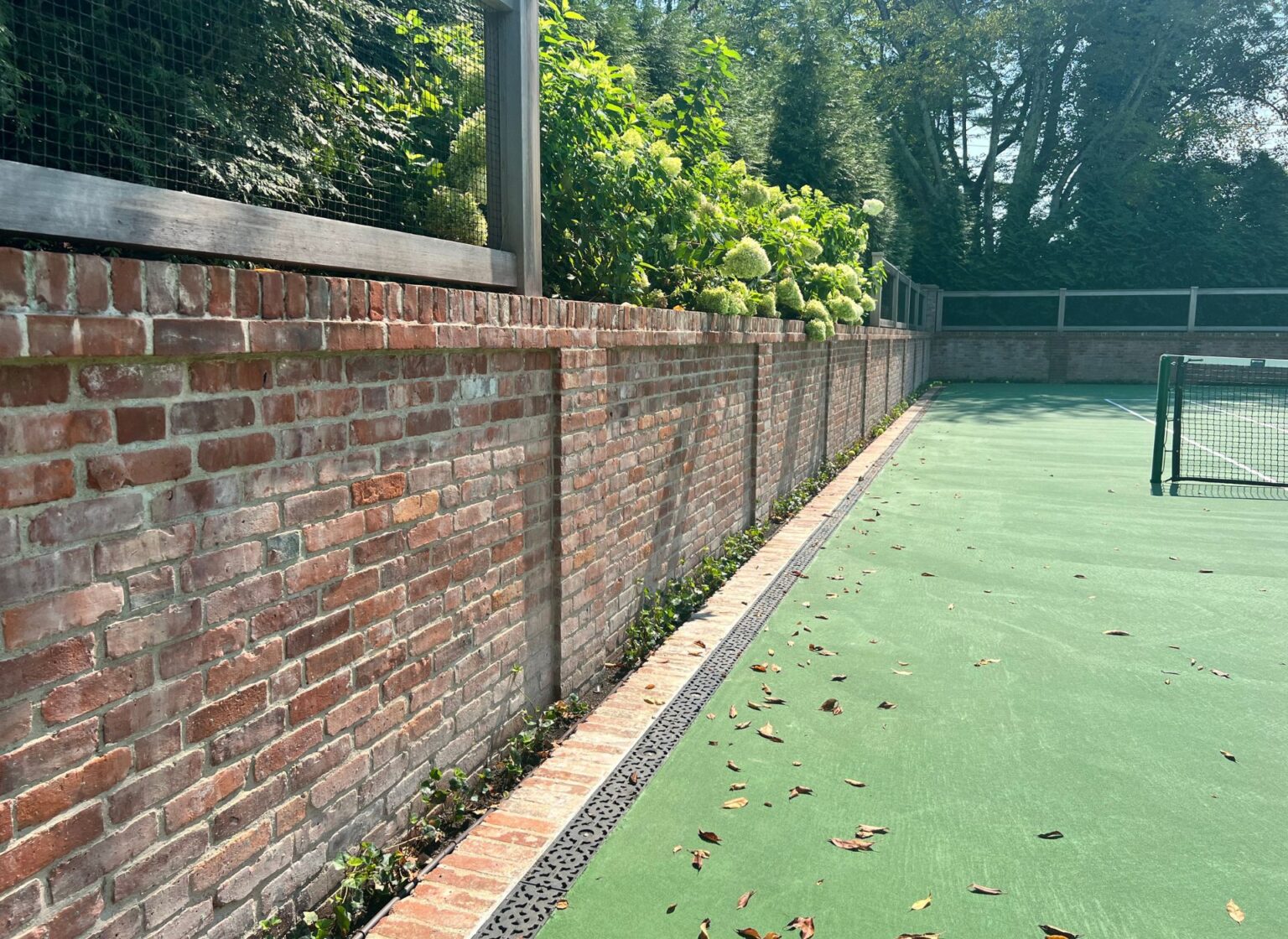 A reclaimed brick landscape wall running along the edge of a tennis court, bordered by fencing and mature trees.