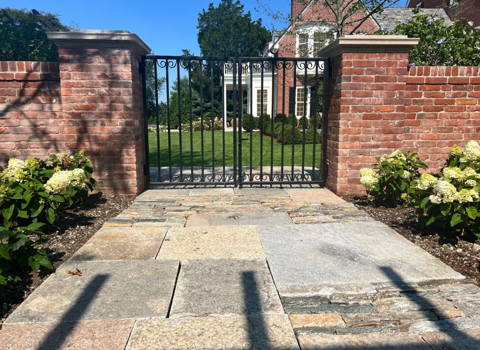 A reclaimed brick perimeter wall with brick piers and a black metal gate, forming an entry to a landscaped property, with a stone walkway leading up to the gate.