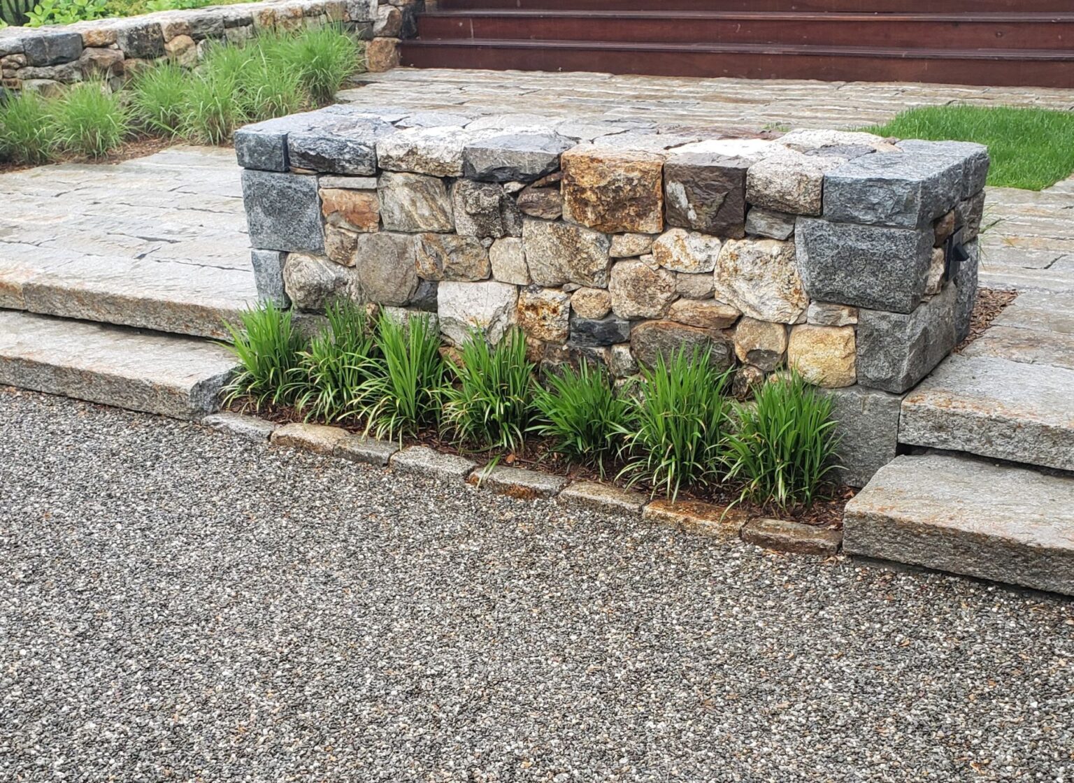 A set of reclaimed granite steps leading up to a patio area, with a low reclaimed stone wall beside the steps and plantings at the base, set in a landscaped yard.