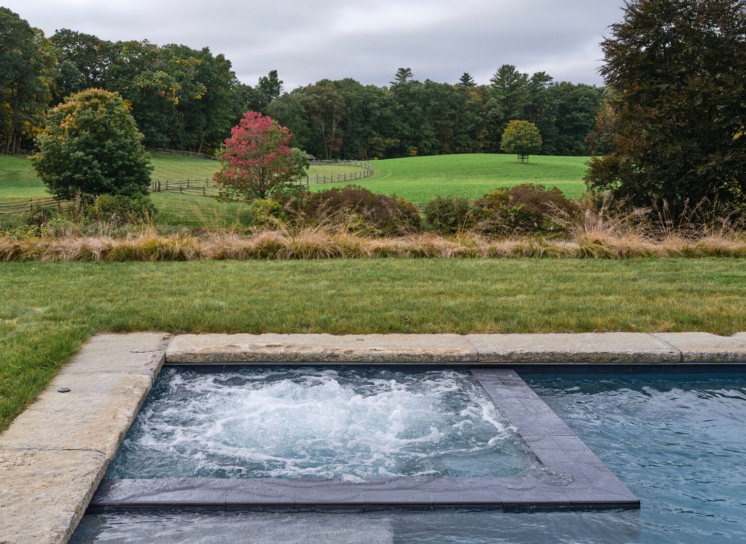 A rectangular swimming pool with a raised spa section finished in reclaimed granite curbing pool coping, featuring thick, weathered stone edges and set against a grassy lawn overlooking rolling fields and trees.