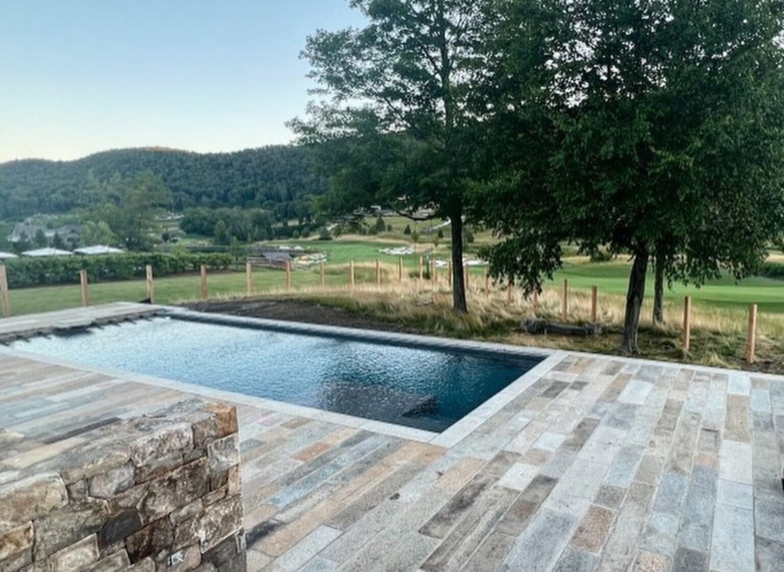 A rectangular swimming pool finished with reclaimed granite plank pool coping, surrounded by matching stone paving and overlooking a pastoral landscape with trees and rolling fields.