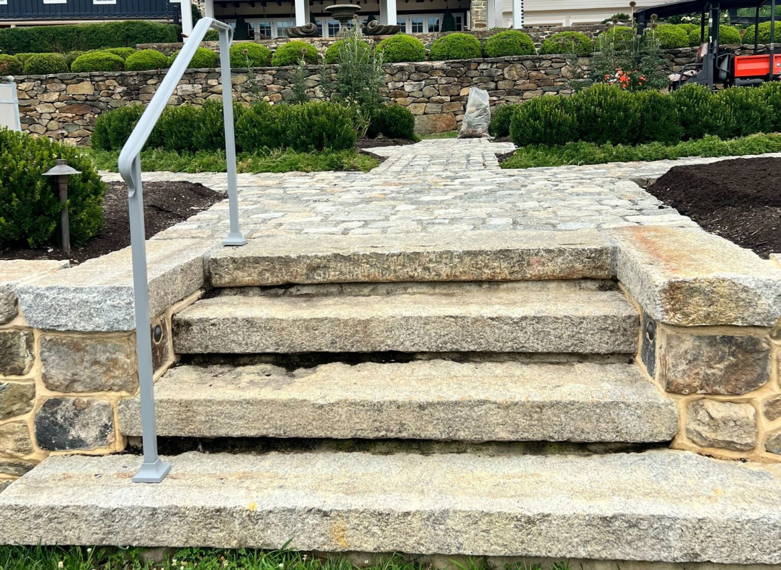 A set of outdoor steps built from reclaimed granite treads, leading up to a stone walkway surrounded by landscaped plantings and a retaining wall.