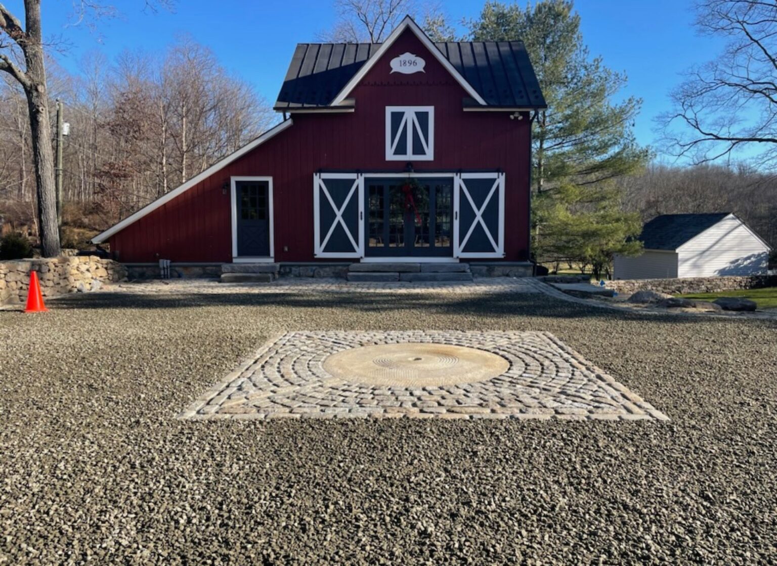A reclaimed antique millstone set as a centerpiece within a gravel driveway, surrounded by a square cobblestone inlay, positioned in front of a red barn‑style building.