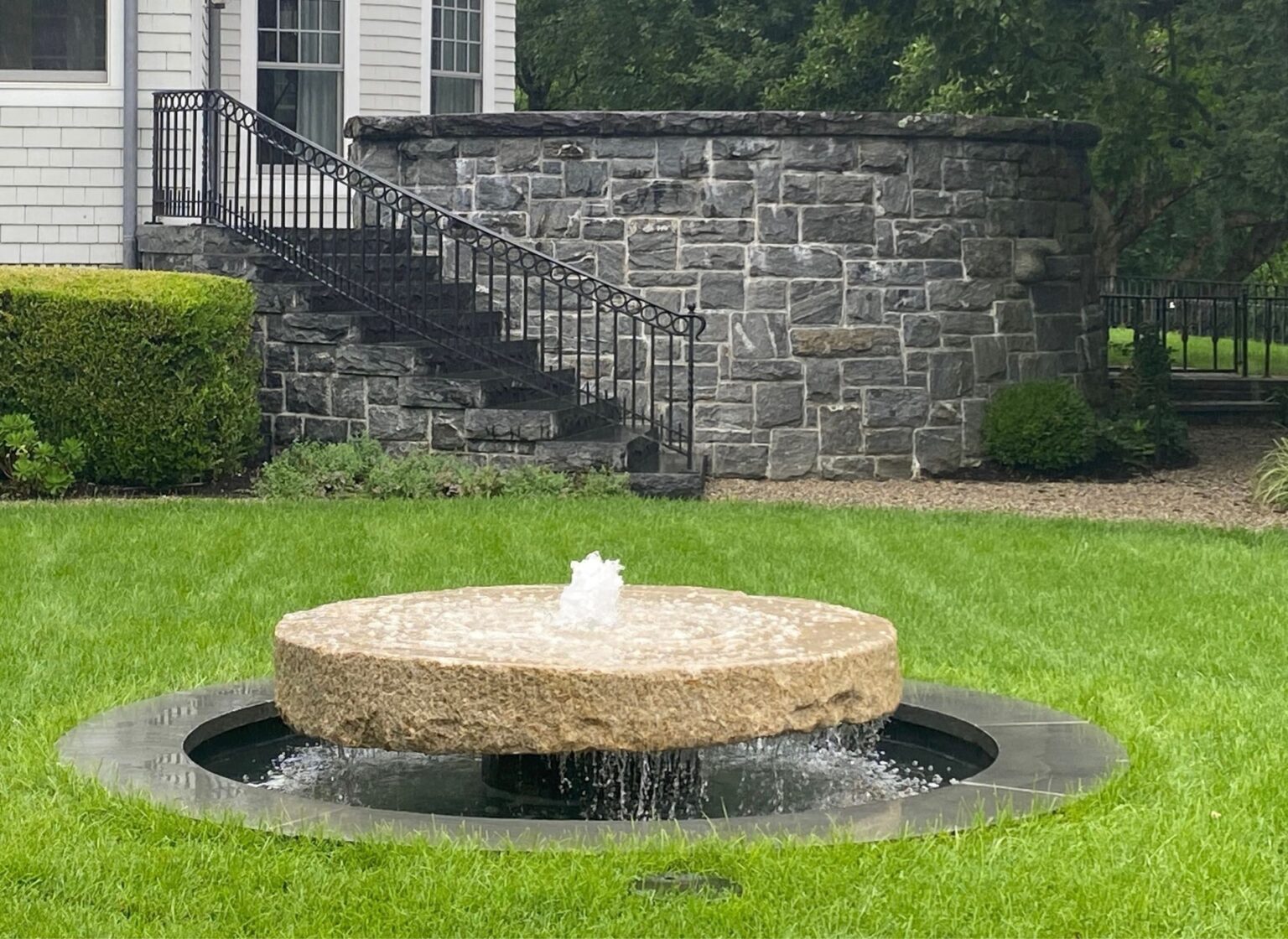 A reclaimed antique millstone fountain set in a manicured lawn, with water flowing from a circular stone millstone into a shallow basin, positioned in front of a stone retaining wall and garden plantings.