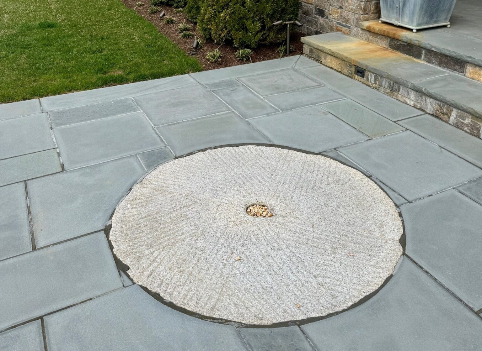 A reclaimed antique millstone set flush within an antique stone walkway, with a circular weathered stone millstone inset among rectangular stone pavers near a set of stone steps.
