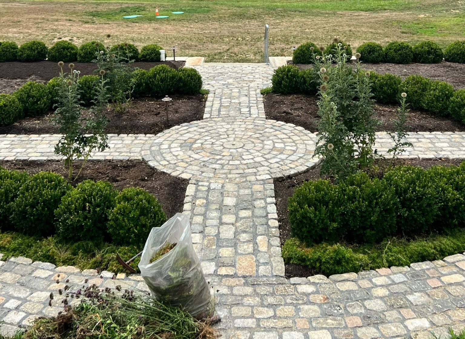 a rustic hardscape design featuring a millstone set as the focal point of a cobblestone circle, with cobblestone paths leading away in four directions