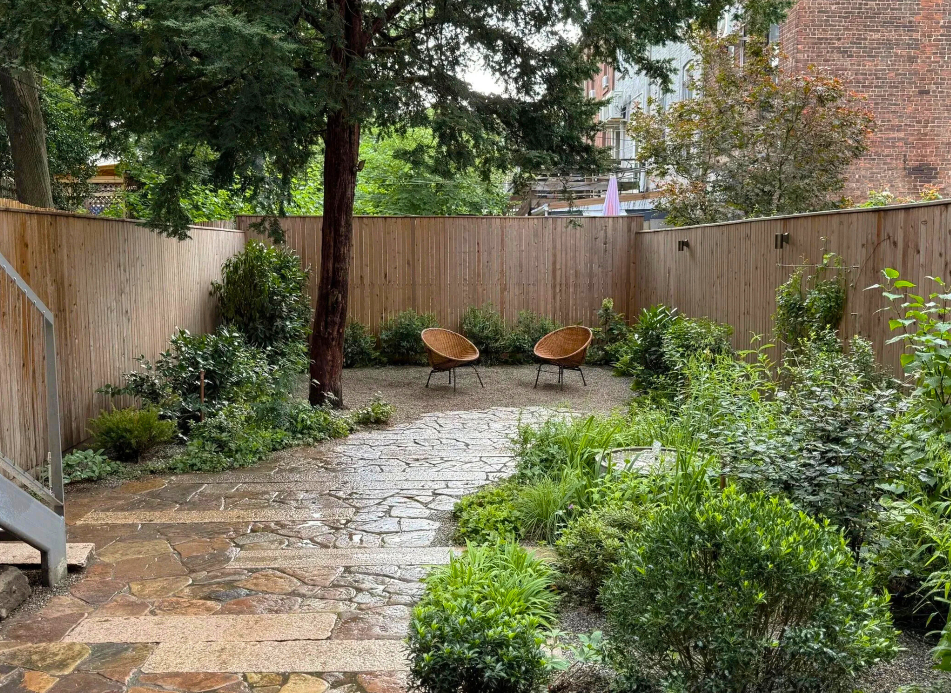 An enclosed urban patio paved with granite planks mixed with irregular fieldstone, forming a winding garden pathway surrounded by dense plantings and wood fencing.